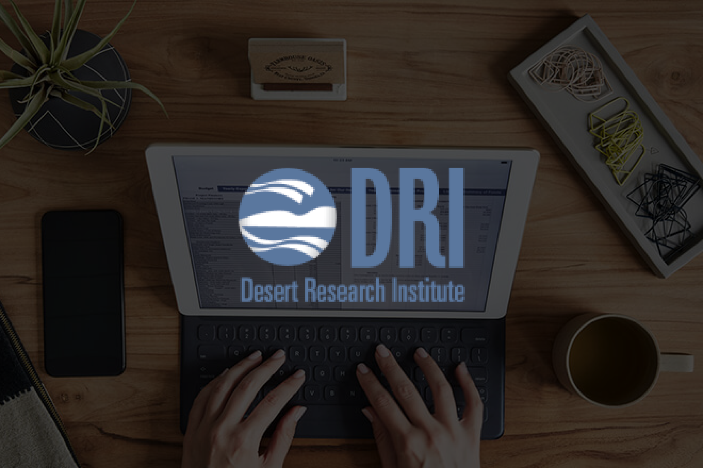 Hands typing on tablet keyboard that is placed on a wooden table with the brand logo overlaid.